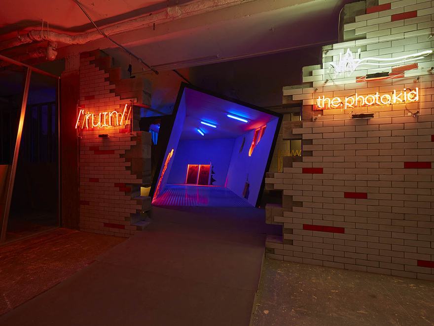 An immersive art installation titled 'Ruin' by Virgil Abloh and Ben Kelly. The scene shows a brick entryway with jagged, broken edges.