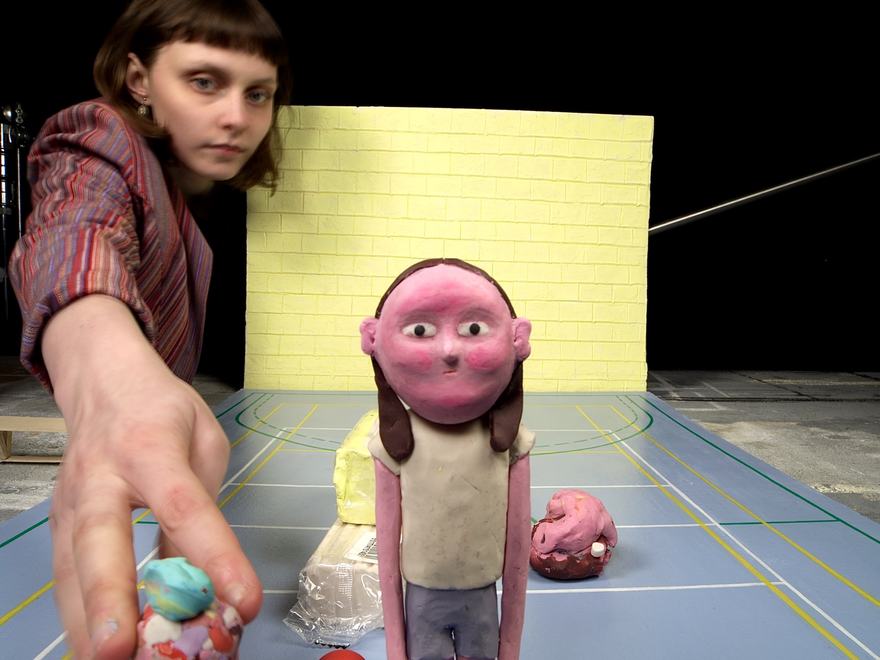 An animator in the back of frame leans towards the camera to manipulate a plasticine character during an animation activity.