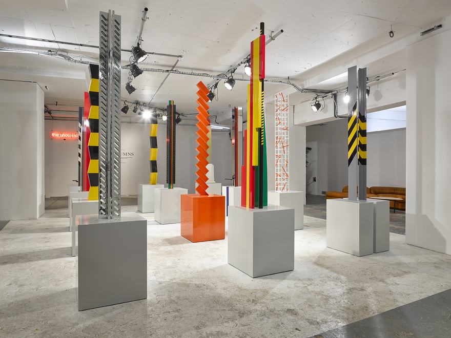 An installation featuring a variety of tall, vertical sculptures presented on minimalist white and orange plinths. Many of the pillars incorporate bold geometric patterns and vibrant colours.