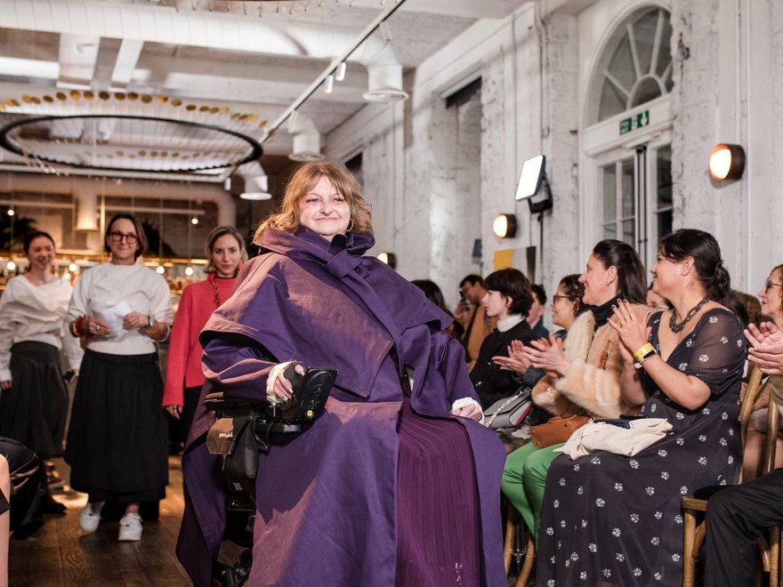 A female wheelchair user models a purple look on a fashion runway with the crowd applausing