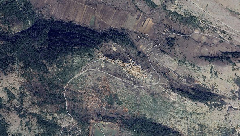 An aerial satellite view of a mountainous or hilly landscape featuring winding roads, varying terrain textures including forested areas and open land, and a cluster of buildings or a small village nestled within the topography.