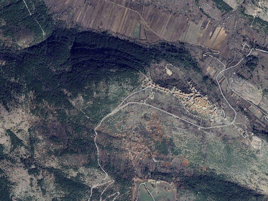 An aerial satellite view of a mountainous or hilly landscape featuring winding roads, varying terrain textures including forested areas and open land, and a cluster of buildings or a small village nestled within the topography.