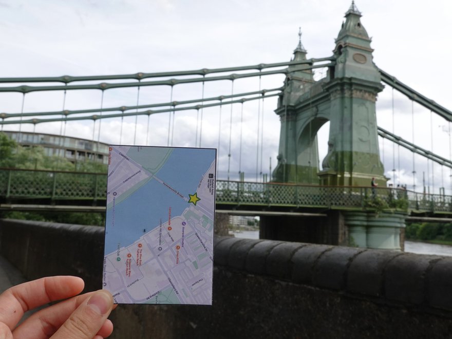 A hand shows a small map in front of Hammersmith Bridge