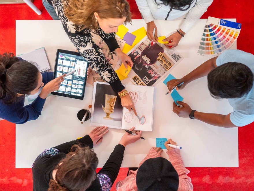A group of students sit around a table, pointing at sketchbooks, colour swatches and digital design references
