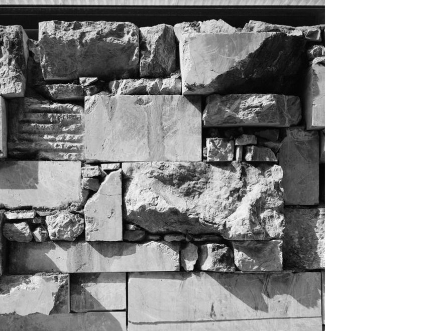 A close-up, black and white photograph of a rustic stone wall constructed from irregularly shaped, rough-hewn stones of various sizes.