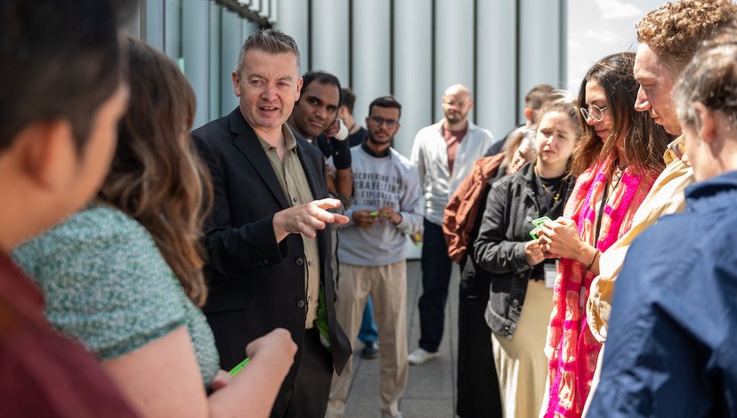 John and the cohort of 2025 Futures Through Design stand out on the RCA Rausing Building balcony, discussing something in the sun.