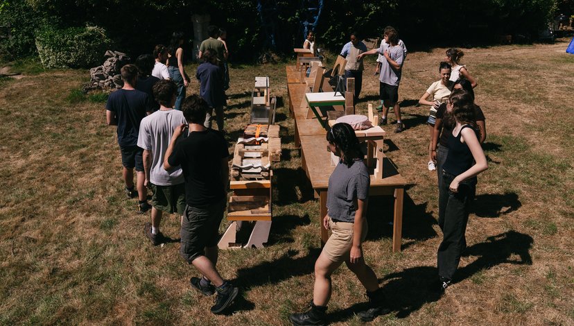 A group of people stand outdoors in a grassy area, gathered around several wooden tables and structures arranged in a long line, possibly displaying items or projects.