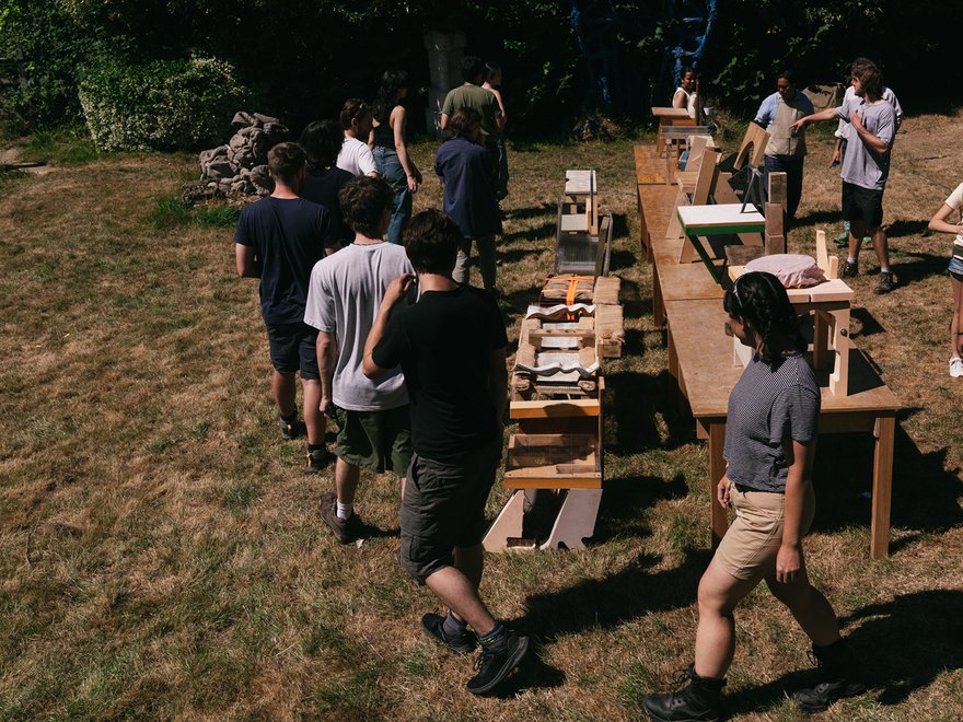 A group of people stand outdoors in a grassy area, gathered around several wooden tables and structures arranged in a long line, possibly displaying items or projects.