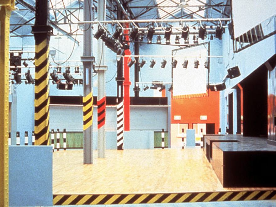 The industrial-themed interior of the legendary Haçienda nightclub in Manchester (FAC51), featuring its signature yellow and black hazard-striped pillars and a large wooden dance floor.
