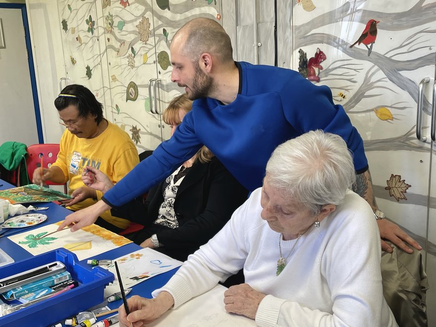Three people sat down painting at a table. Another person is standing and guiding one of the painters.
