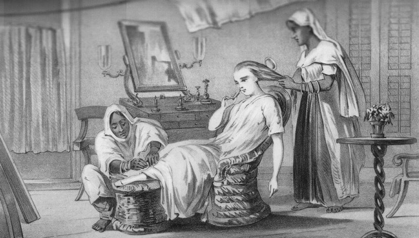 An antique lithograph depicting a woman, likely a magistrate's wife during the British Raj, being attended to by two Indian servants in a domestic setting. One servant brushes her hair while another massages her feet.