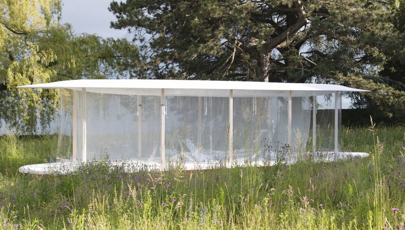 A minimalist, rectangular pavilion with a flat roof and transparent walls stands in a grassy field, surrounded by trees under a bright sky. The structure appears modern and light, blending into the natural outdoor setting.