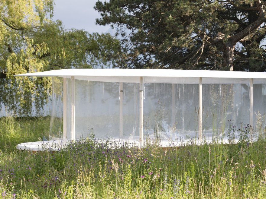 A minimalist, rectangular pavilion with a flat roof and transparent walls stands in a grassy field, surrounded by trees under a bright sky. The structure appears modern and light, blending into the natural outdoor setting.