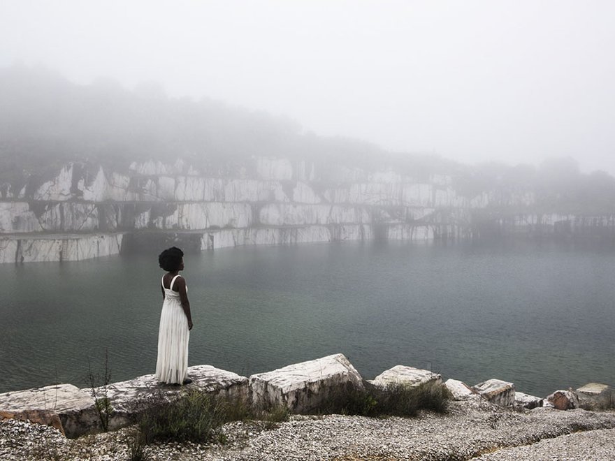 A woman in a white dress stands on the edge of a dramatic bay looking at the sea