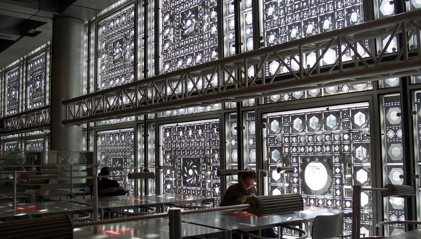 Interior view of the Institut du Monde Arabe (Arab World Institute) in Paris, showing the distinctive geometric patterned facade with diaphragm-like apertures controlling light, and people seated at tables in a library or study area.
