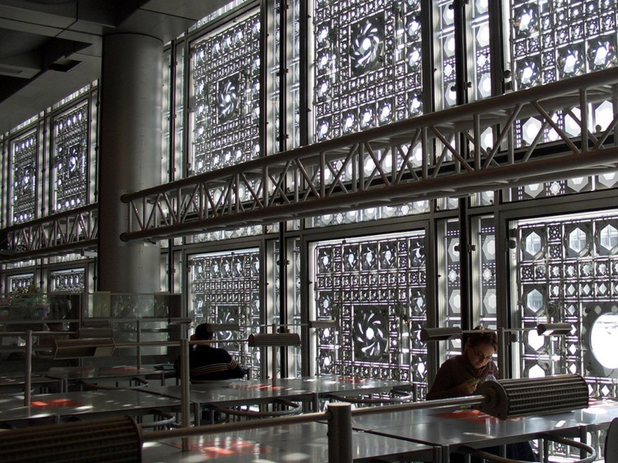 Interior view of the Institut du Monde Arabe (Arab World Institute) in Paris, showing the distinctive geometric patterned facade with diaphragm-like apertures controlling light, and people seated at tables in a library or study area.
