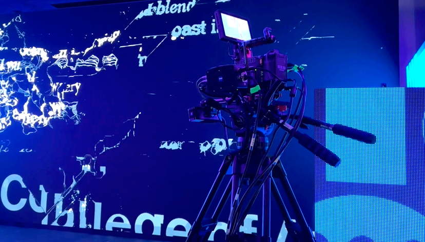 Professional video camera on tripod filming in front of a vibrant digital screen with abstract text and graphics.