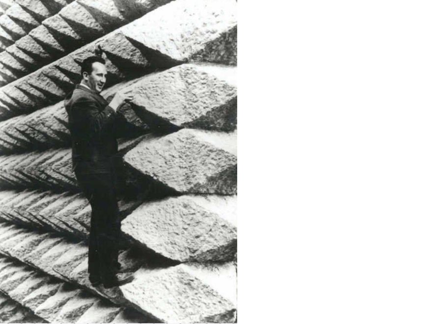 A black and white photo of a man in a suit standing on a massive, textured surface resembling a woven pattern or a stack of large, diamond-shaped blocks, possibly a close-up of a dam or a large rope structure, with the man looking towards the camera.