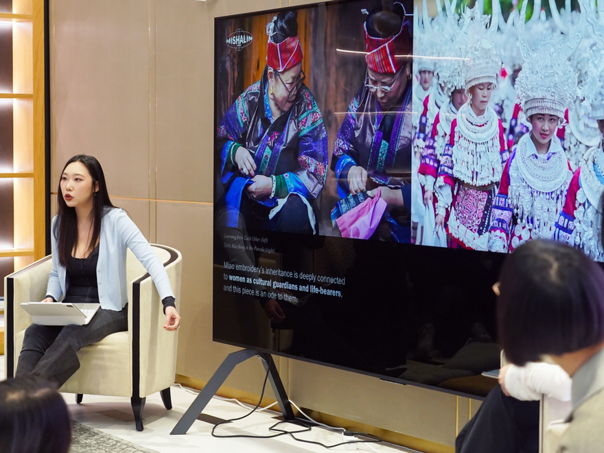 A woman gives a talk with a large screen behind showing women in traditional dress