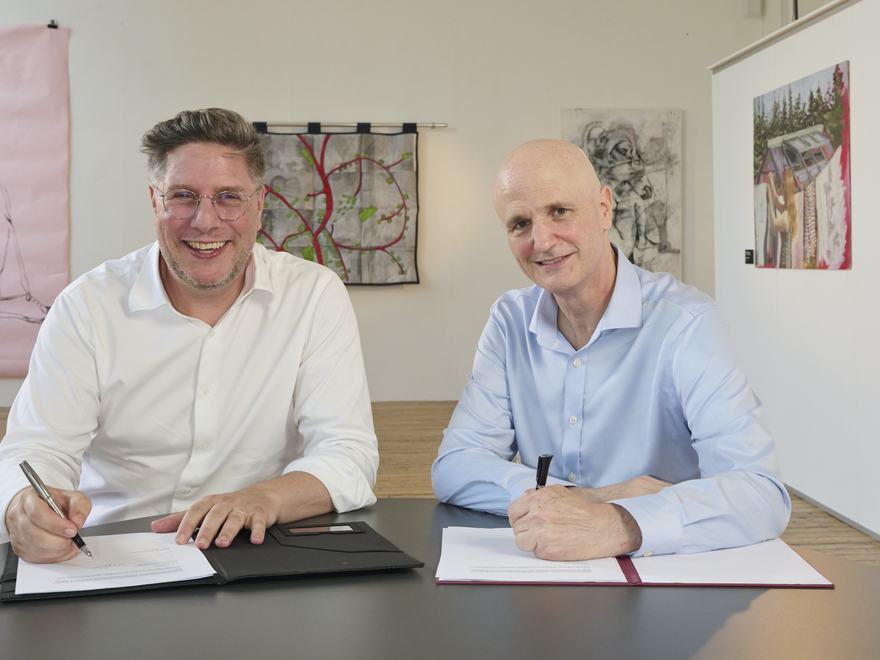 Two men sit behind a desk holding pens above document papers, as if to sign an agreement together. They look at the camera and behind them is a room with colourful art visible on the walls.