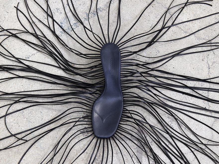 Black sandal with long, thin leather cords fanning outward on a concrete surface.