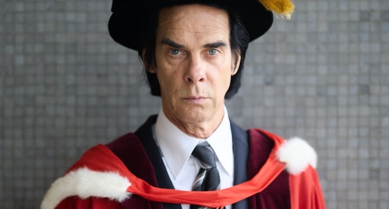 A portrait of musician Nick Cave in academic regalia, featuring a black cap, a white shirt with a striped tie, a dark suit, and a red and maroon robe.