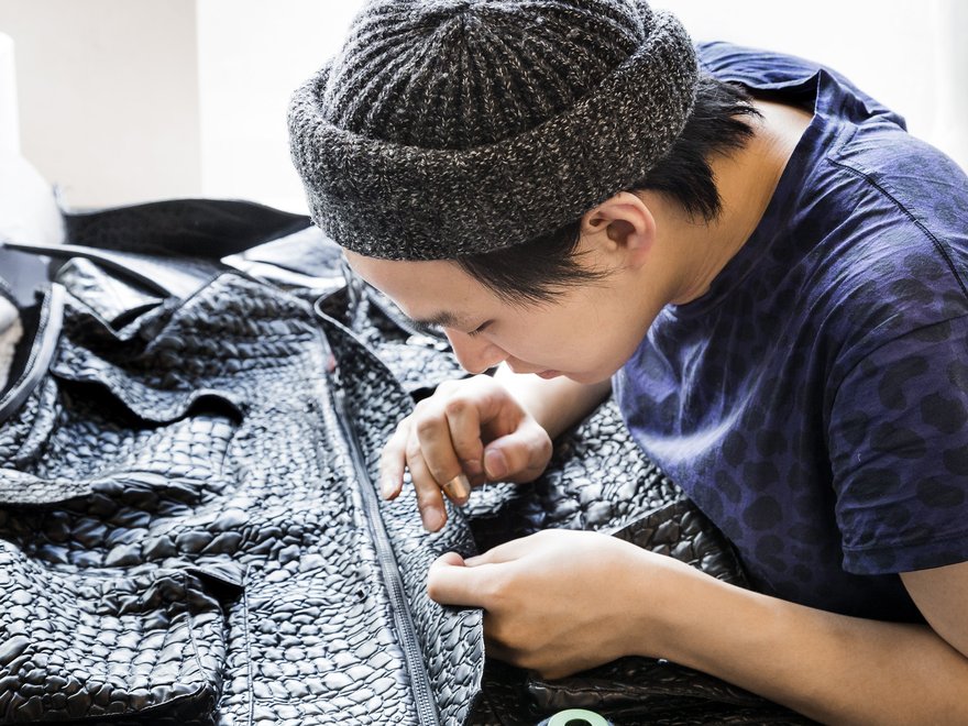 A student in a knitted hat hand sews a garment made of black faux crocodile leather
