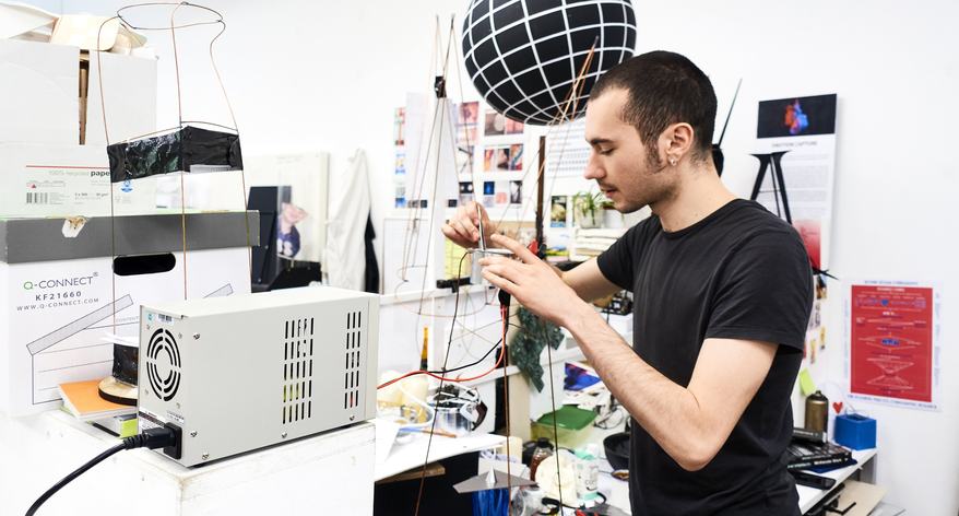A man in a black t-shirt working with electronic equipment and wires in a workshop, featuring a large black-and-white checked sphere suspended above his desk.