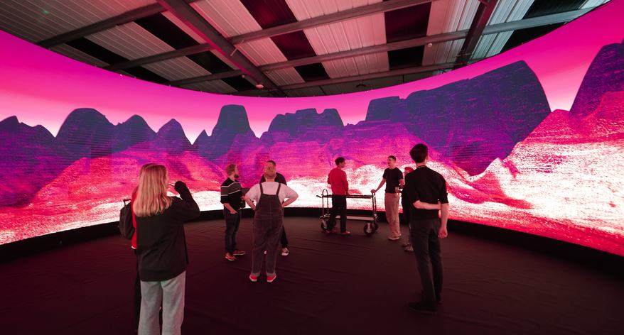 Students in an immersive multimedia facility at Norwich University of the Arts. A wraparound screen displays a digitally illustrated landscape in purple, pink and black.