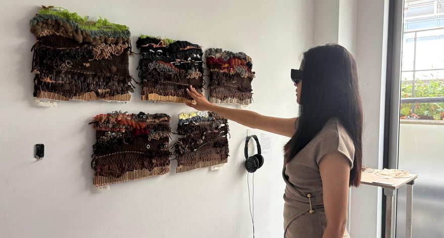 A person wearing blacked out sunglasses holds out their hand to feel a textured work hung on the wall