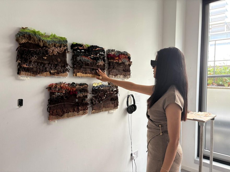 A person wearing blacked out sunglasses holds out their hand to feel a textured work hung on the wall