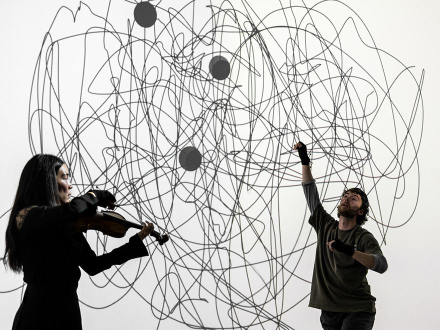 A violinist plays beside a performer reaching upward, framed against a large digital projection of tangled black scribbles and dark circles.