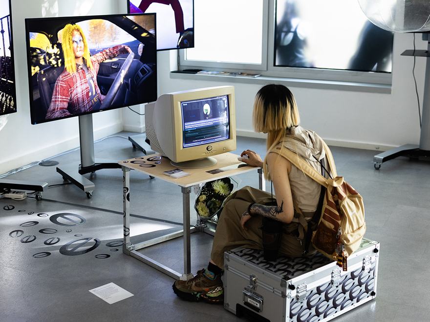 A visitor with blonde-highlighted hair interacts with a vintage CRT computer monitor displaying digital artwork by Xiaoyao Ma, set alongside a larger flat-screen display at RCA2026.