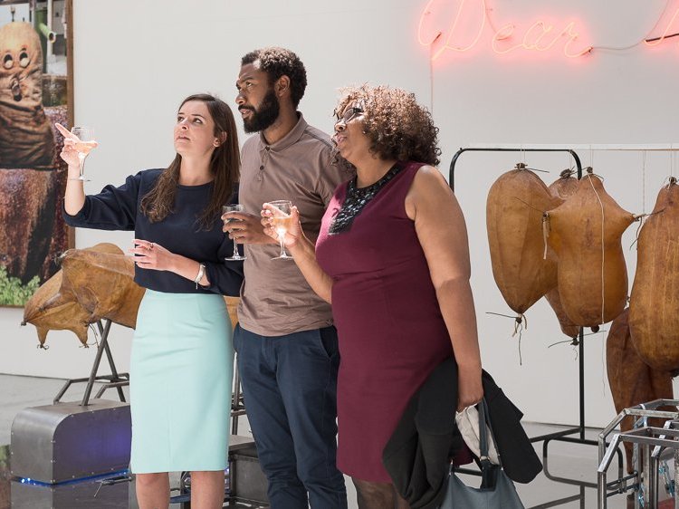 Three people discuss works in an exhibition. Neon text and inflated leather sculptures hand on the rear wall.