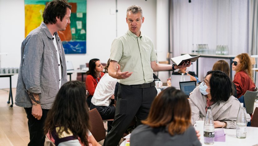 A male instructor in a light green shirt leads a workshop, gesturing with his hands while holding a notebook. He is surrounded by several participants seated at a white table in a bright, modern classroom setting.