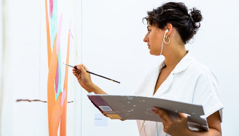 An image of a woman wearing headphones, painting a canvas that hangs on a wall.