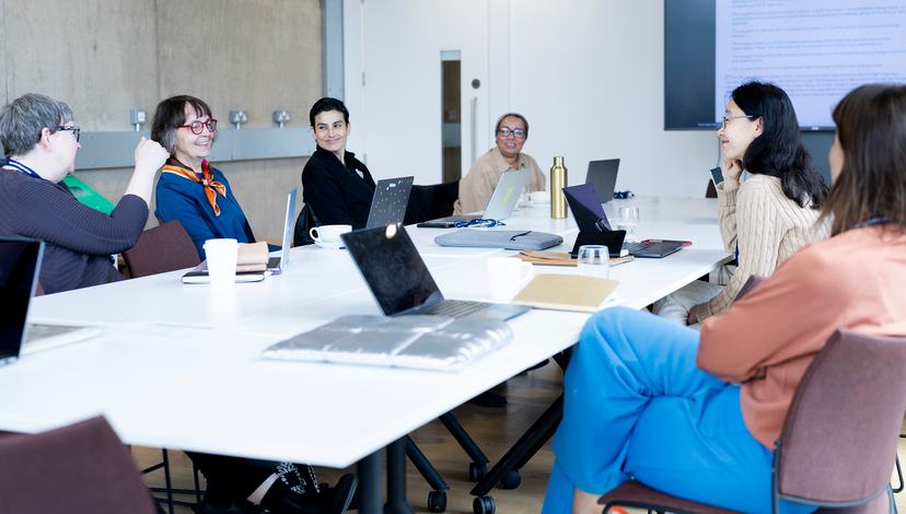 A diverse group of people sitting around a long white conference table in a modern meeting room, engaged in a discussion with laptops and notebooks open.