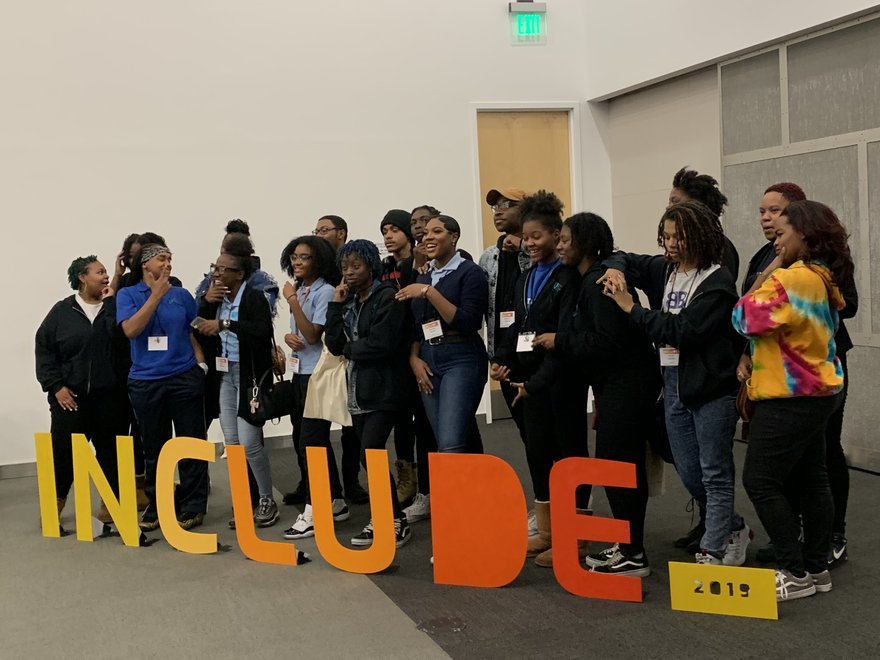 A group of 20 black participants at the Include 2019 conference, standng in front of large cut out letters spelling 'Include'.