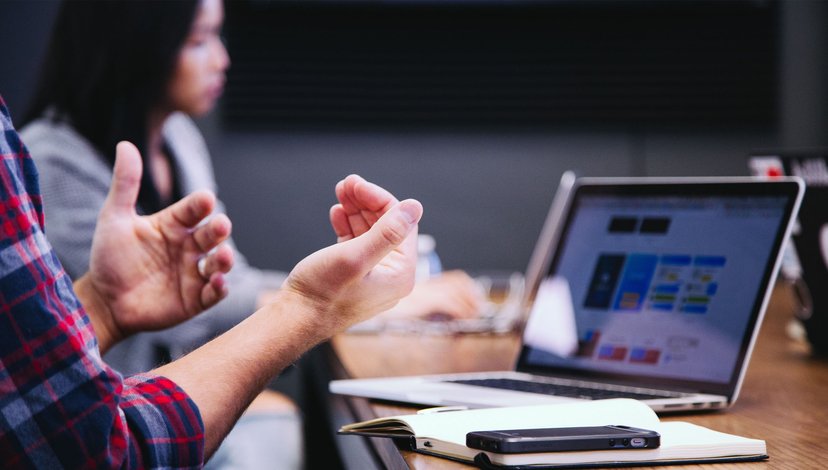 Two people in a meeting, one gesturing while speaking, with laptops and a phone on the table.