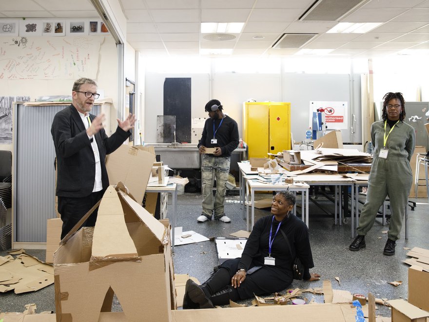 Course leader standing and gesturing while standing next to a cardboard model in a creative workshop space, with three participants observing and engaging.
