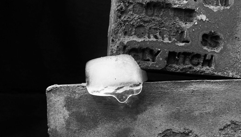 A close-up, black and white photo of a single melting ice cube resting on a rough stone surface, with harshly carved text visible on a weathered tombstone or monument behind it.
