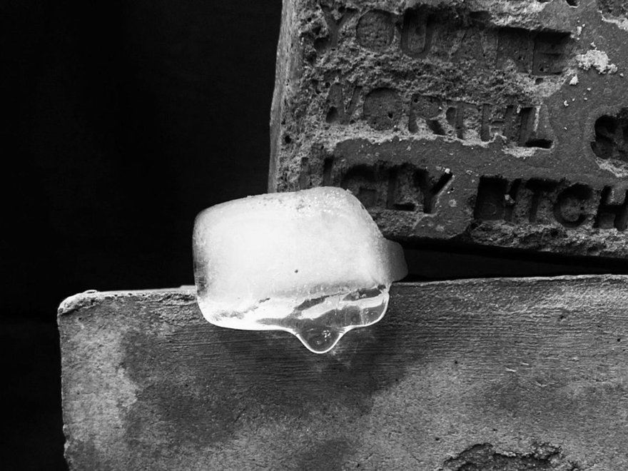 A close-up, black and white photo of a single melting ice cube resting on a rough stone surface, with harshly carved text visible on a weathered tombstone or monument behind it.
