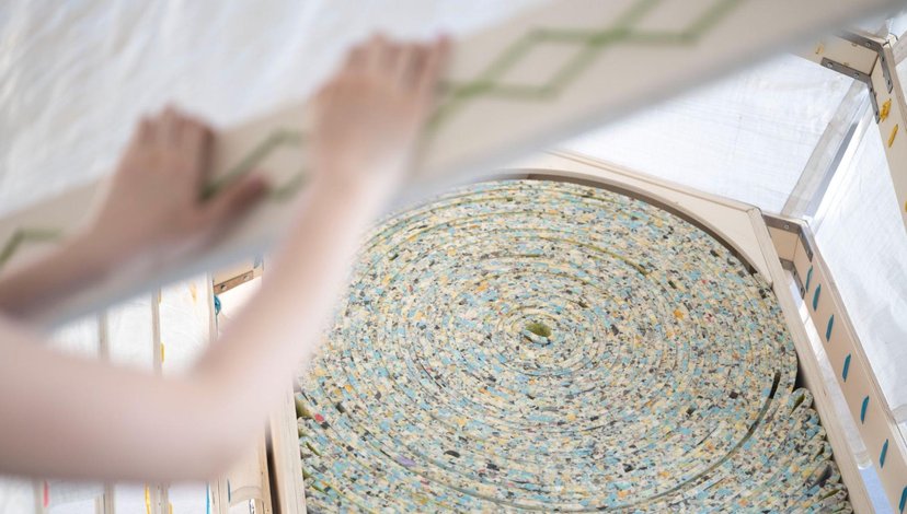A close-up of a person installing a lid or cover over a circular coil of multi-colored, speckled foam padding that fills a wooden hexagonal container.