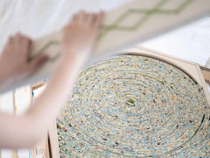 A close-up of a person installing a lid or cover over a circular coil of multi-colored, speckled foam padding that fills a wooden hexagonal container.