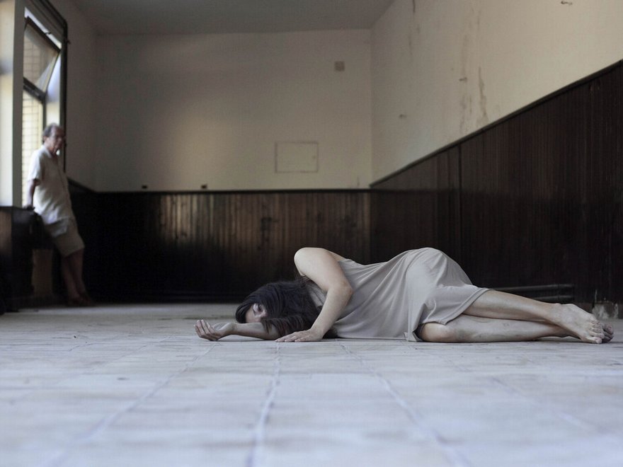 A photo of a woman lying on the ground inside a room, a man leans against a window in the far left corner