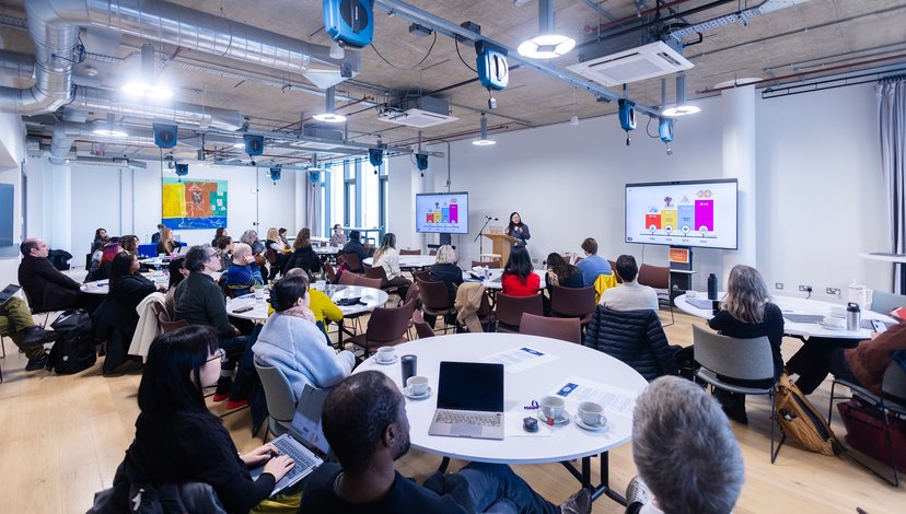 A group of people sitting at round tables in a modern, industrial-style presentation room, listening to a woman speaking at a podium. Large screens on the wall display colourful bar charts and data visualisations.