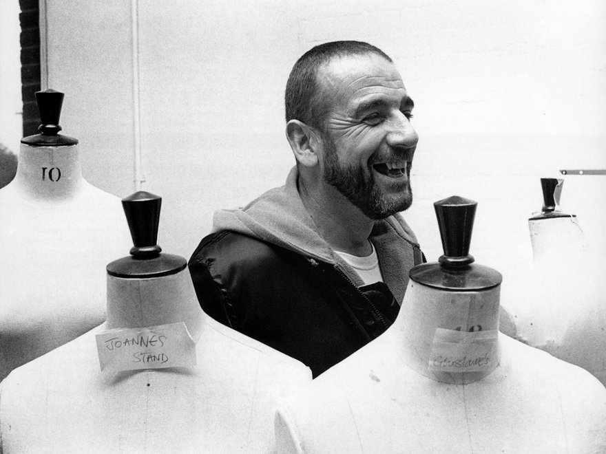 A black and white photograph shows a smiling John Miles in the centre of the frame, surrounded by four fashion design mannequins.