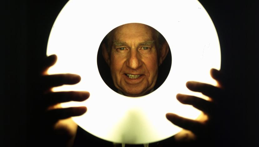 A man holding a light in front of his face, with his hands visible in the foreground. The background is dark, creating a mysterious atmosphere.