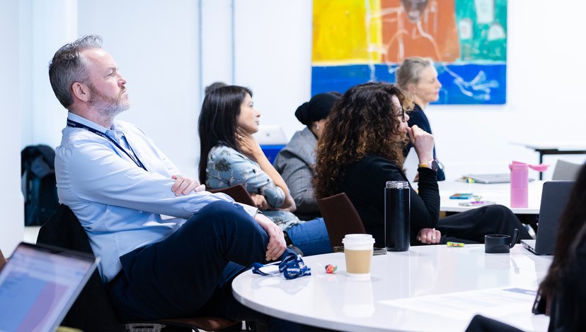 A diverse group of people sitting around a white circular table in a brightly lit meeting room, attentively listening to a presentation.