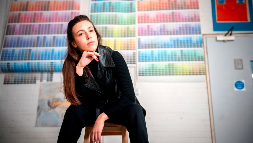 A young woman sitting on a stool in a studio with large colour charts on the wall.